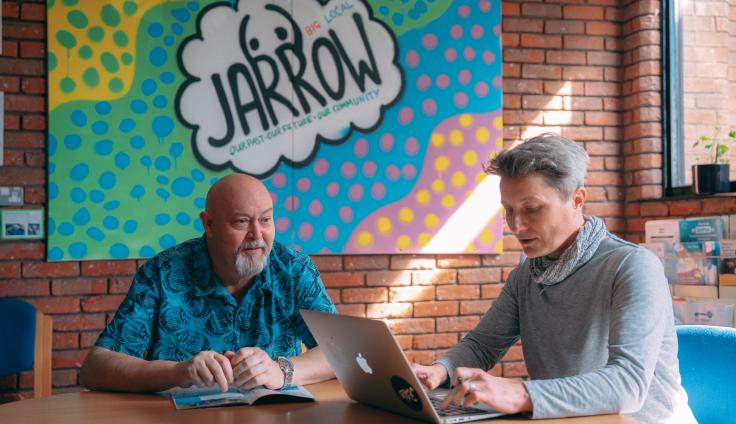 Two people at a desk, looking at a laptop, with a 'Jarrow' mural on the wall behind them.