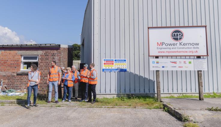A group of people in hi vis jackets stand next to a large grey building with a sign reading 'MPower Kernow'