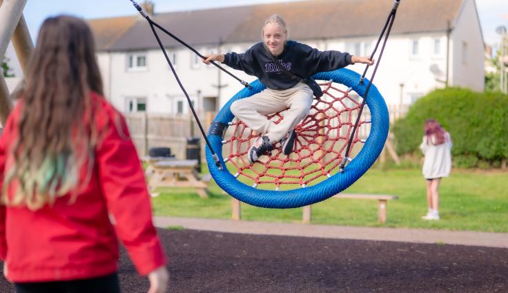 A young person sitting on round blue swing smiles directly at the camera.
