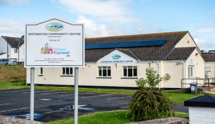 A sign reads 'Distington Community Centre, home of Distington Big Local'. Behind is a car park and a cream coloured community building.
