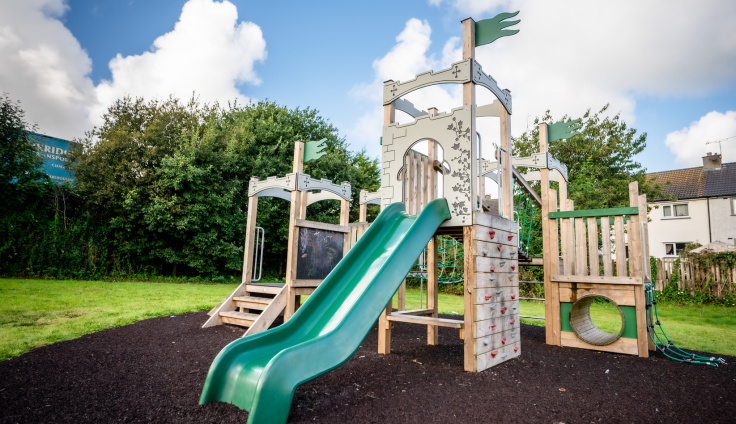 A new wooden playground with green slide, green flags and steps, set up in a landscaped park in front of a row of houses.