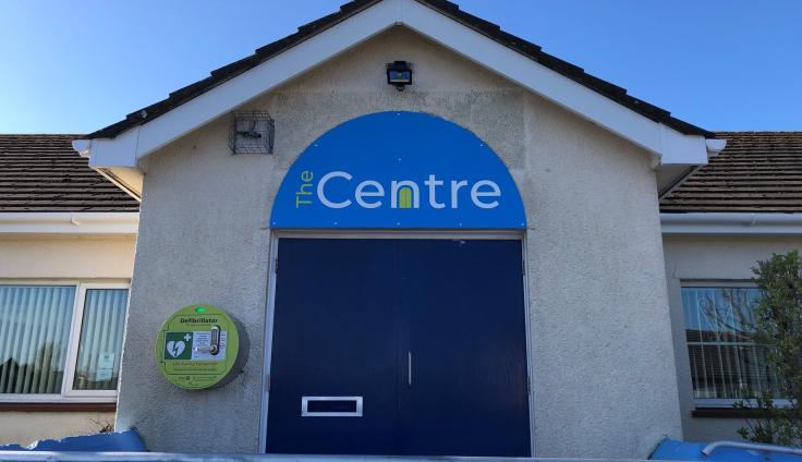 Building with navy doors and 'The Centre' logo in a semi-circle along with a defibrillator attached to the wall in Ewanrigg, Cumbria.