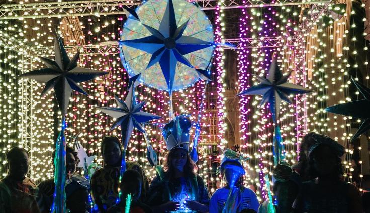 A crowd of people are silhouetted against strings of pink and yellow lights as they hold up giant stars on sticks.