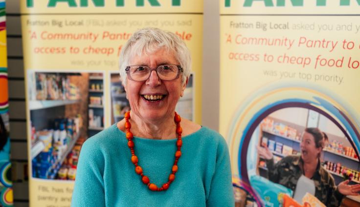 A cheerful older woman with short grey hair and glasses smiles warmly at the camera. She's wearing a bright turquoise sweater and a vibrant orange beaded necklace. Behind her is a yellow community information banner about the "Community Pantry" initiative, with visible text mentioning access to affordable food locally. The banner includes photographs showing shelves stocked with food items, illustrating the community pantry in action.