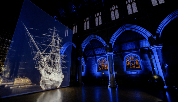 A white ship is projected in light against a dark blue screen in a church lit with blue light. It is nighttime.