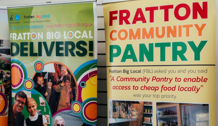 Two colourful community information banners are displayed side by side. The left banner features vibrant circular designs and community photos, with text reading "FRATTON BIG LOCAL DELIVERS!". The right banner has a yellow background with bold text reading "FRATTON COMMUNITY PANTRY" in red, orange, and green letters. Below this, it explains that Fratton Big Local asked the community what they wanted, and their top priority was "A Community Pantry to enable access to cheap food locally." The bottom of the right banner shows photographs of what appears to be the community pantry in operation, displaying shelves stocked with food items.
