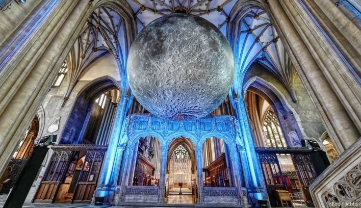 A giant lifelike moon hangs in the centre of a gothic church nave, lit by blue lights from below.