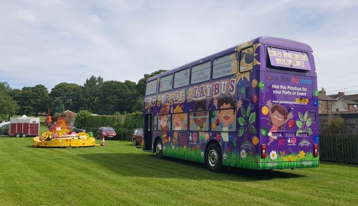 A purple bus parked in a recreation field, covered in colourful cartoons and the writing Big Purple Playbus.