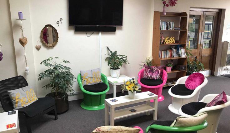 A room with bookcases and a media wall, and a seating area with colourful tub chairs, a table and a vase of flowers.