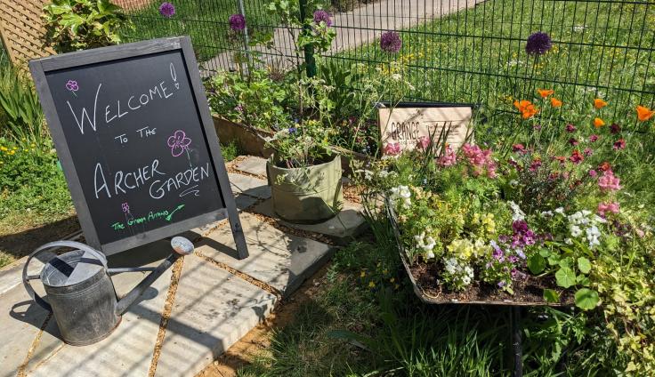 An outdoor setting. A black sandwich-board style handwritten sign reads 'Welcome! To the Archer Garden'. A watering can sits next to the sign on a paved path, and both are surrounded by wooden planters and other boxes full of colourful flowers.