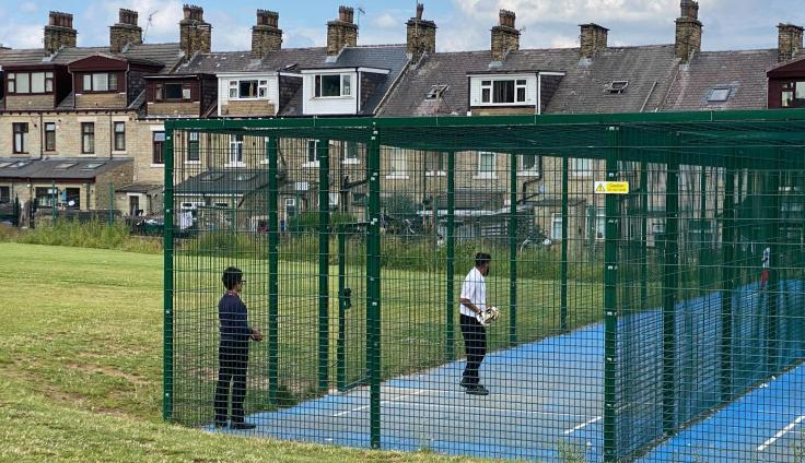 Two people playing a game in a green steel-framed play area with a blue floor, set in a grassed area adjacent to houses.