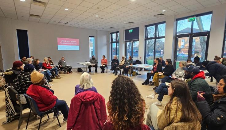 A large group of people sitting on chairs set out in a circle, in a bright room with large windows.
