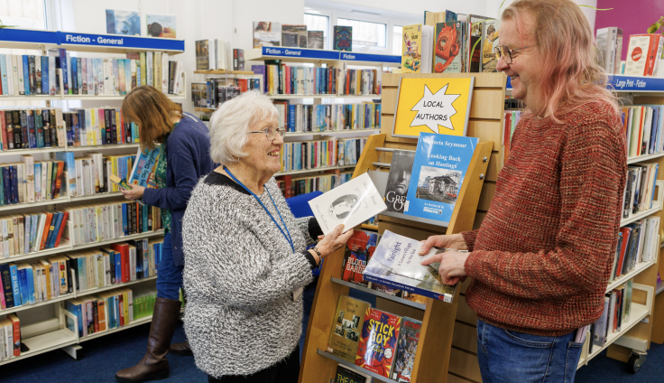Interior of a library showing a woman and a man engaged in conversation near a wooden book display stand marked 'LOCAL AUTHORS' with a yellow sign. The woman on the left is elderly with white hair, glasses, and wearing a gray cardigan with a blue lanyard, holding a book. The man on the right has shoulder-length blonde hair, glasses, and is wearing a red knitted jumper, also holding a book. Behind them are extensive bookshelves with blue section markers reading 'Fiction - General' and 'Large Print - Fiction.' Another person can be seen browsing books in the background.