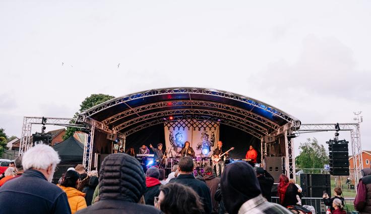 A group of people standing looking towards musicians performing on a covered stage.