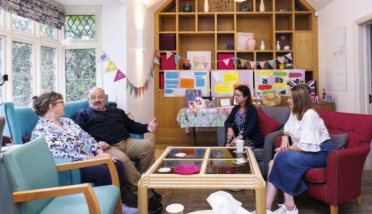 Three women and a man sit in conversation in a common room. There is wooden shelving behind them, a window to the left, and colourful bunting strewn behind them. In the foreground there is a paper coffee cup and a box of tissues on a table.