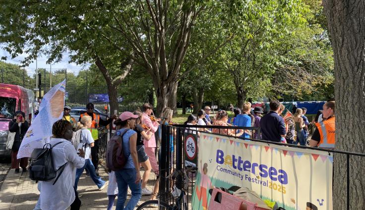 A group of people walking between black railings where there is a sign reading 'Battersea community festival 2025'.