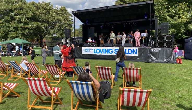 Young people perform on musical instruments on stage at Battersea Community Festival. Two people are dancing in front of the stage, other people are sitting on striped deck chairs facing towards the stage.