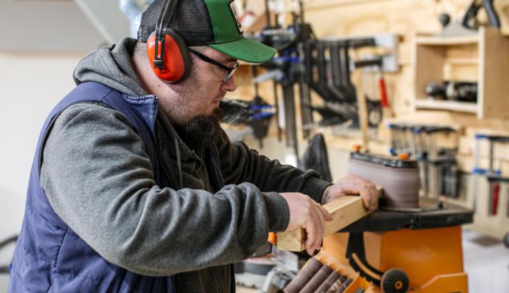 A white man wearing ear protection and a peak-cap, working on a piece of timber using a belt-sander, with lots of tools hanging on a tool board in the background.
