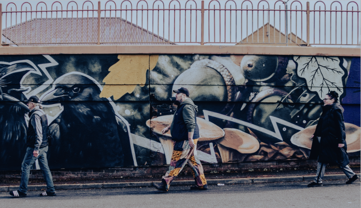 Three people walk past a large street art mural on a wall beneath a metal fence with decorative loops. The detailed graffiti features a prominent black raven or crow on the left side, along with crows, acorns, mushrooms, against a dark blue background. From left to right, the pedestrians include two men wearing caps and black coats. One of them is wearing colourful patterned trousers. Behind them is a woman in a long dark coat. All captured mid-stride as they pass the colourful urban artwork.