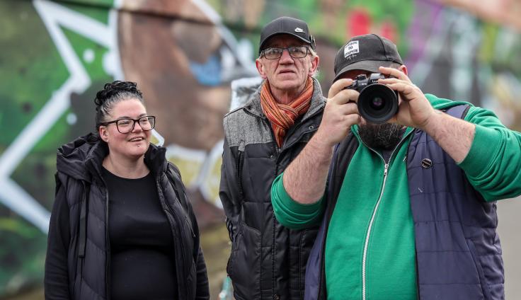 Three people stand together outdoors, with colorful graffiti-covered walls in the background. On the left, a young woman with glasses and dark hair in a bun wears a black hoodie and smiles at the camera. In the center, an older man wearing glasses, a black cap, and a striped scarf over a gray and black jacket looks toward the camera. On the right, a bearded man in a green shirt and dark vest holds a large camera up to his face, actively taking a photograph.