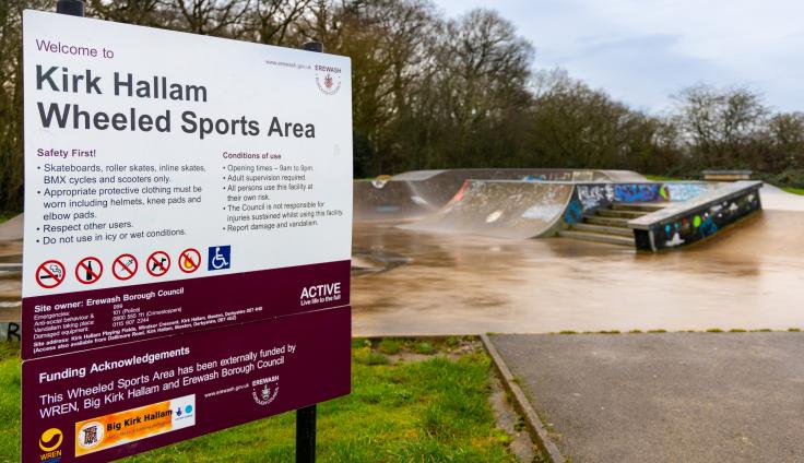 A sign reading 'Kirk Hallam Wheeled Sports Area' with a concrete ramps in the background