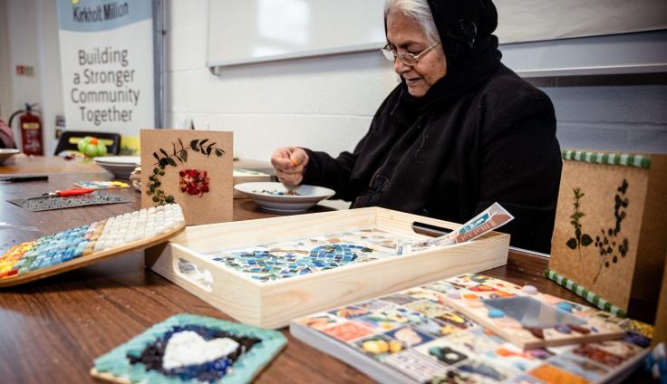 A woman sitting at a table making a small glass mosaic.