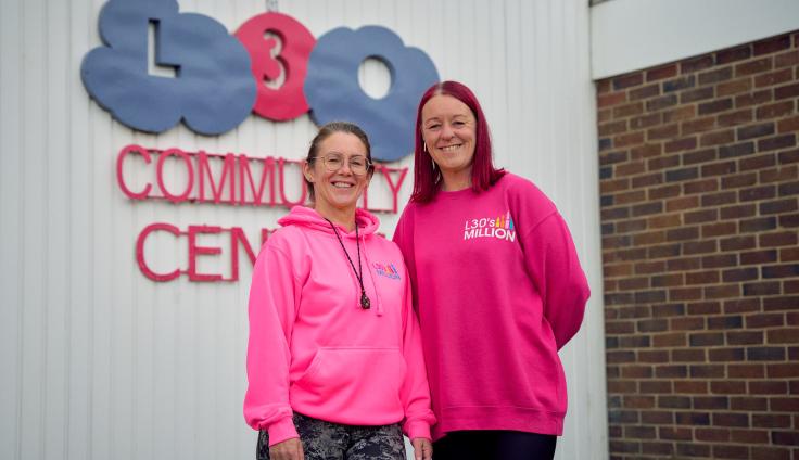 Two women smiling at the camera wearing bright pink jumpers, in front of a sign reading 'L30 Community Centre.'
