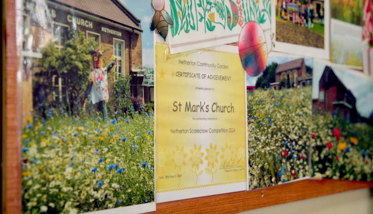 A noticeboard with a yellow certificate for St Mark's Church surrounded by wildflowers in a green space.