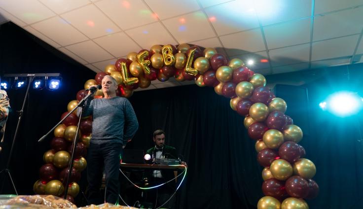 A person on a stage in front of a balloon arch, with the letters LFBL, speaking into a microphone.