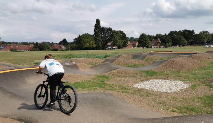 An undulating concrete bike track with bunkers on a sandy, grassy area with trees in the background and a boy riding on a bike.