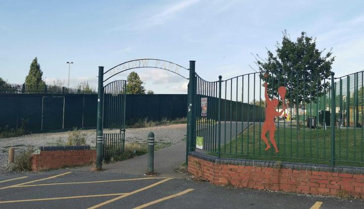 A metal archway marks the entrance to a park with the words "Meadow Park" in the ironwork. An outline of a woman throwing a tennis ball in the air to serve is attached to the railings. A concrete path leads into the park. A tree and blue cloudy sky can be seen in the background.