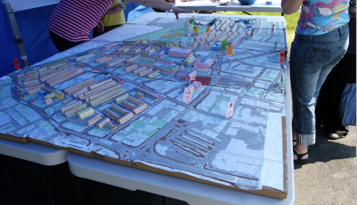 A giant 3D map of housing plans is set out across two trestle tables, surrounded by three people leaning over it. Their waist upwards is cropped. They are standing under a blue gazebo.