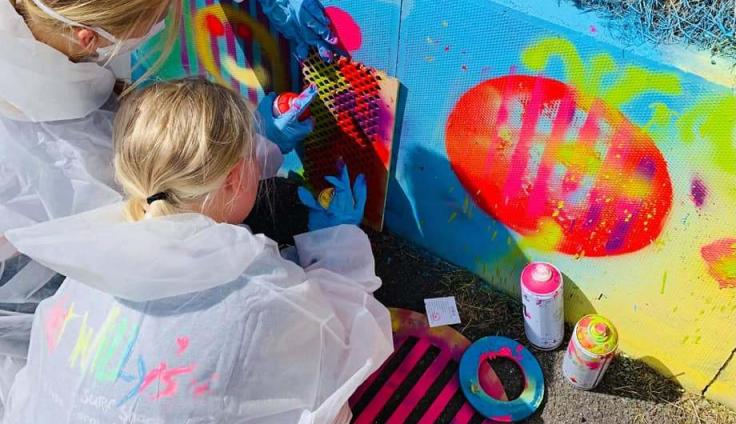 Two people with blonde hair wearing protective clothing stencilling on a painted blue wall.