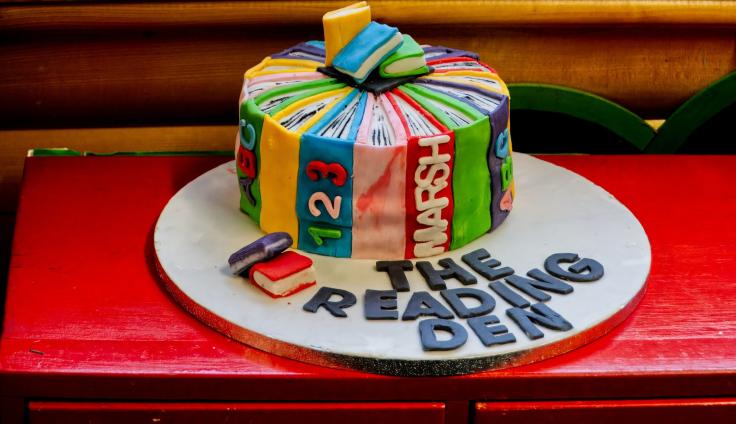 A colourful cake iced with book designs with icing letters spelling out 'The Reading Den'.