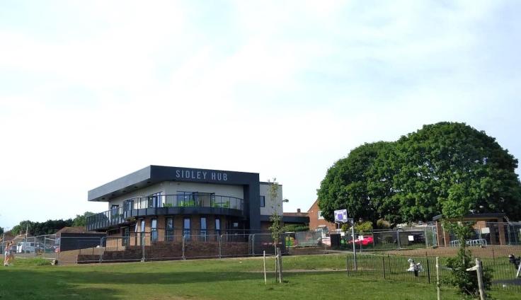 A two-storey brick building with a balcony and writing reading 'Sidley Hub' and green space in the foreground.