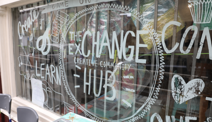 A storefront window displays hand-painted white lettering and decorative designs on glass. The main text reads "THE EXCHANGE" with "CREATIVE COMMUNITY" and "HUB" prominently featured. The design incorporates circular motifs with decorative zigzag borders and heart shapes.