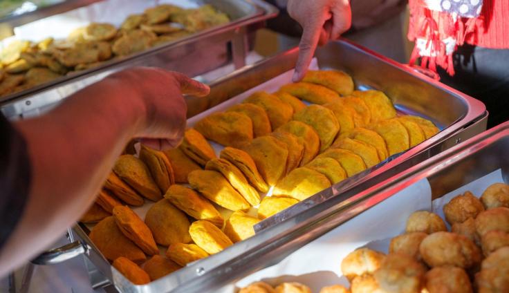 Steel warming dishes filled with pastries and fried food, with two people pointing at food items.