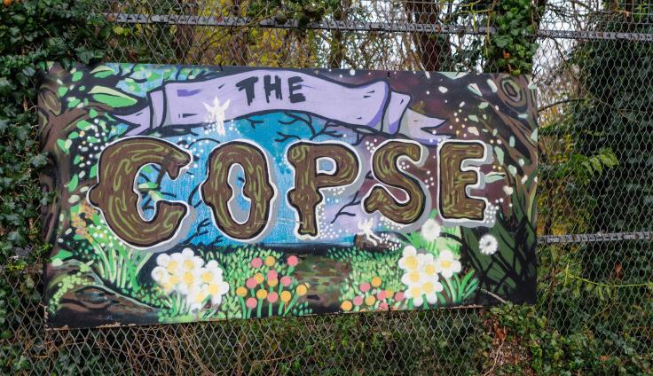 A woodland area with a colourful sign on railings reading 'The Copse.'
