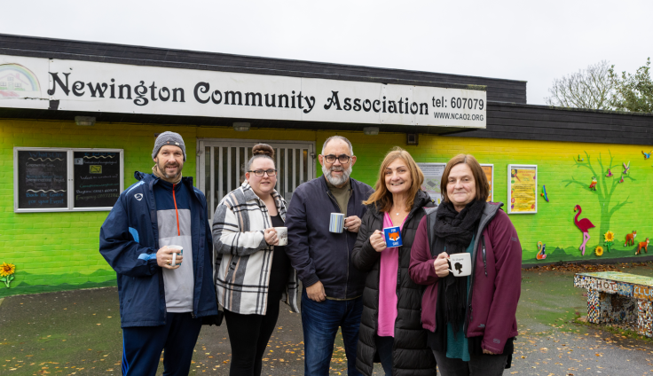 Five people stand holding mugs and smiling outside Newington Community Association, a green painted low-rise building with noticeboards, a painted tree, flowers, and birds. From left to right, a man with a beard wears a grey beanie hat, navy blue coat, jumper and sports trousers; a woman with glasses and a bun on top of her heads with black leggings, a black top and a white, black and grey tartan coat; a man with grey hair, beard and moustache, black glasses wears blue jeans, a navy jacket and a green thsirt; a woman with mid-length red hair has a pink scarf, a long black puffa jacket; a woman with short brown hair has a black scarf, a burgundy jacket and a green and black dress.