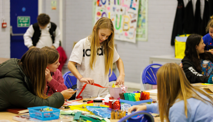 A teenage girl in a white tshirt and long blond hair looks down at a craft table and holds a red ribbon. Three other girls surround her and look at her, with other colourful craft materials including pens and paper on the wooden table. There is a teenage boy in the background wearing a white and black top, standing in front of a blue fire escape door. On the white-washed brick wall behind them is a large poster that reads "Chill Club" in colourful letters. Two more teenage girls can just be seen on the right of the frame.