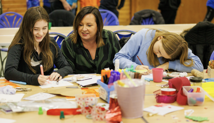 At a crafts table full of pens and different art materials, two young people sit drawing with a woman called Nova, a local artist. The table is long with blue chairs, and there is another empty table behind them.