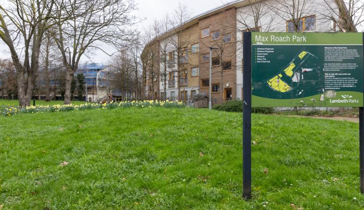 A black sign reads Max Roach Park surrounded by grass and trees and a three-storey building in the background