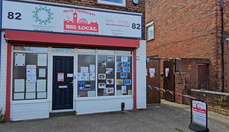 A white cladded store front, with a sign above showing the North Cleethorpes Big Local logo and the windows are filled with printed photos and community notices.