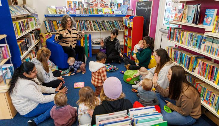 A group of women and young children surrounded by bookcases, watching a woman tell a story.