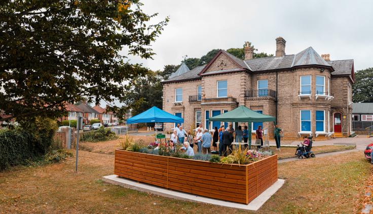 A wooden seating area with parasols in front of a large Victorian building.