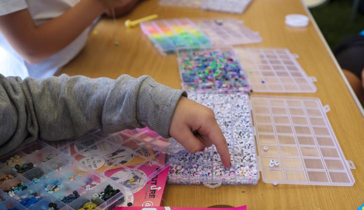 A close-up on a child's arm in a grey jumper, pointing at some beads in a tray. There are other trays of colourful beads and sequins on the wooden table, and a glimpse of another child in a white tshirt crafting at the table.