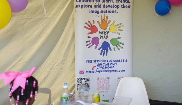 A close-up view of a 'Messy Play' promotional banner displayed at what appears to be a children's activity event. The banner features colourful handprints arranged in a circle around the text 'MESSY PLAY' and describes sessions for children to learn, create, explore and develop their imaginations. Contact details are provided. QR codes are visible at the bottom of the banner. In the foreground, there are water bottles, supplies, and a white plastic chair. Colourful balloons are visible in the background, and there's a glimpse of a little girl wearing a pink bow and pink top on the left side of the image.