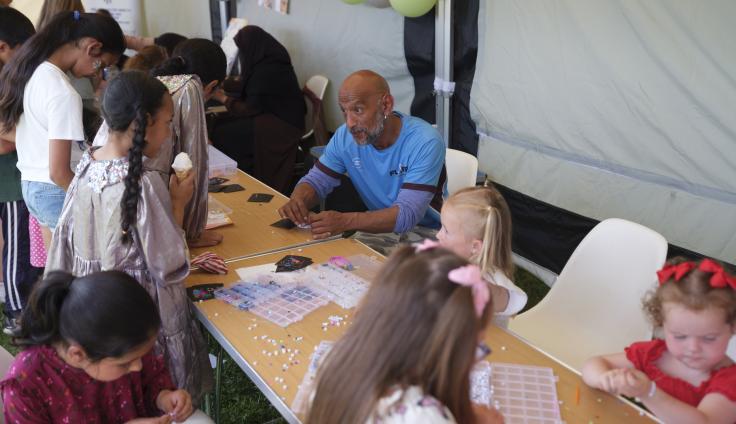 A community craft activity taking place outdoors under a white tent or canopy. A man in a light blue t-shirt sits at a wooden table surrounded by children of various ages who are engaged in what appears to be a puzzle or craft project. The table is scattered with small white pieces, papers, and craft materials. Children are standing and sitting around the table, actively participating in the hands-on activity.