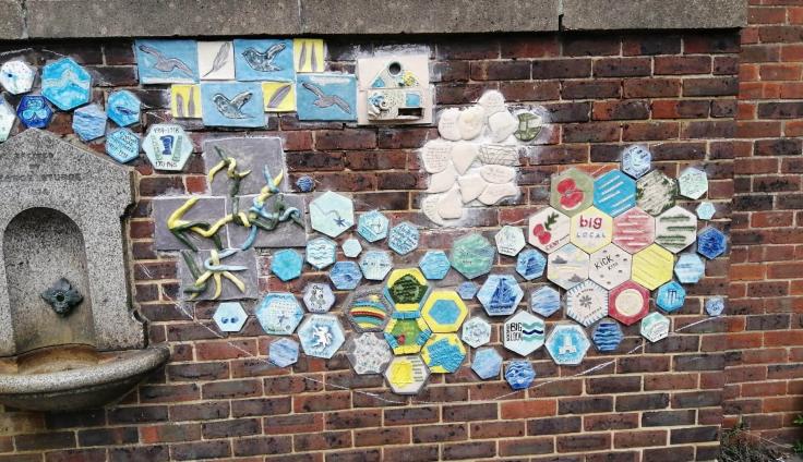 Colourful hexagonal tiles in shades of blue and yellow with drawings of red poppies and birds on a brick wall next to a drinking fountain.