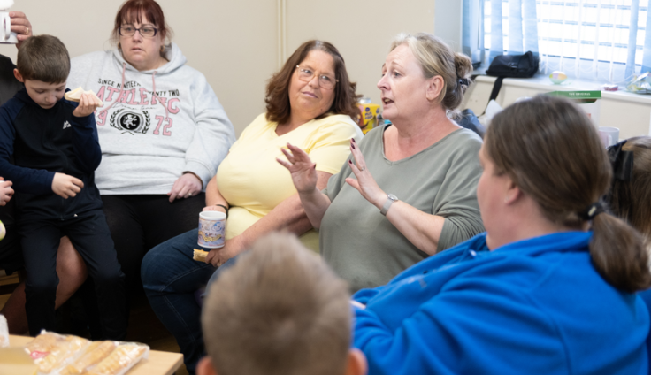 A woman in a green t-shirt has her hands up an in front of her and is speaking to a circle of people. Next to her sits a woman wearing a yellow tshirt holding a cup of tea. A woman in blue with brown hair has her back to the camera and there is a glimpse of the back of a boy's head. A woman in a grey hoodie and a little boy in black also sit in the circle.